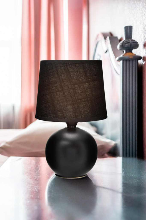 A lamp with a black ball ceramic base and black fabric shade, sitting on a bedside table.
