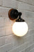Black metal wall light with opal shade and natural wood mounting block on white brick background