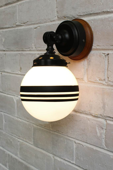 Black wall light with natural wood mounting block and opal shade with three stripes against white brick background