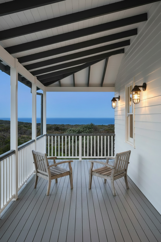 A coastal porch with black ceiling beams, two wooden chairs, two wall lights and ocean view in background