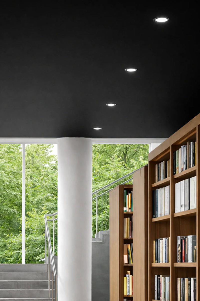 Four white LED downlights in a black ceiling of a librabry with large windows and timber bookcases. 
