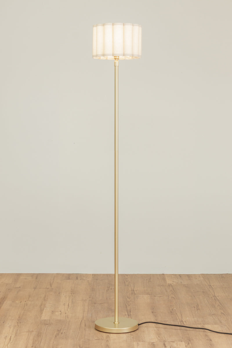 Gold/brass candle stick floor lamp with a fabric lampshade on a timber floor and muted background. 
