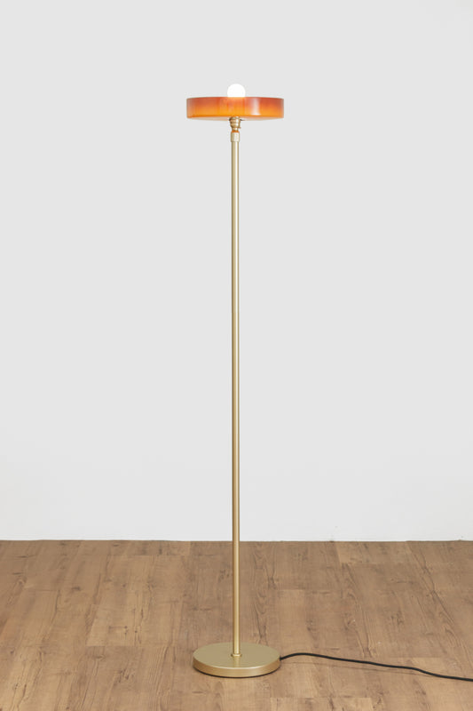 Minimal floor lamp with a tall slim gold/brass stem, circular amber shade and matching round base on a timber floor.