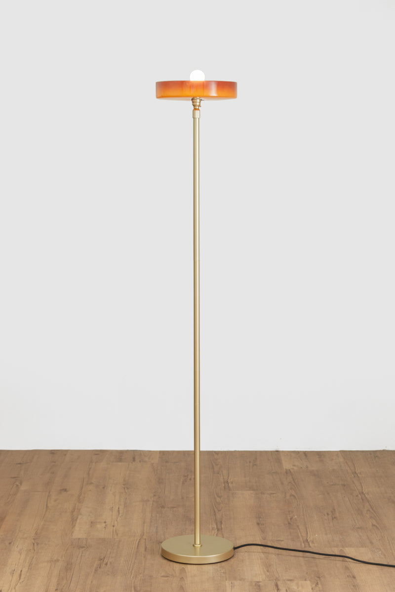 Minimal floor lamp with a tall slim gold/brass stem, circular amber shade and matching round base on a timber floor.