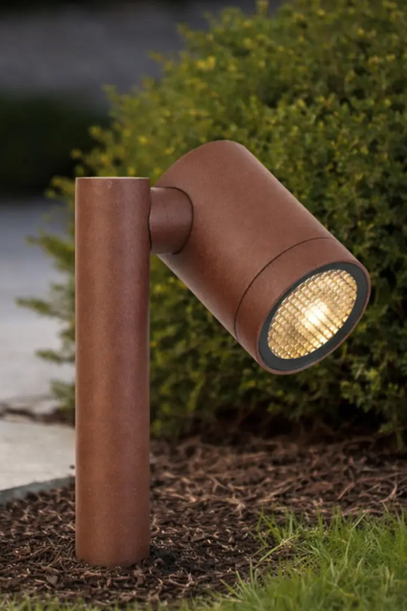 Corten short garden spike light with an adjustable spotlight head, lit in a garden bed in front of a hedge