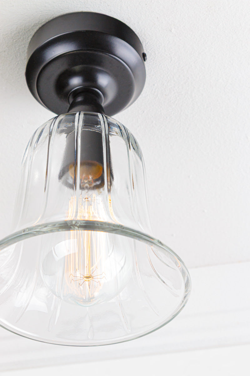 Close up of the black batten holder with a clear glass, bell shaped shade placed on a white ceiling. 