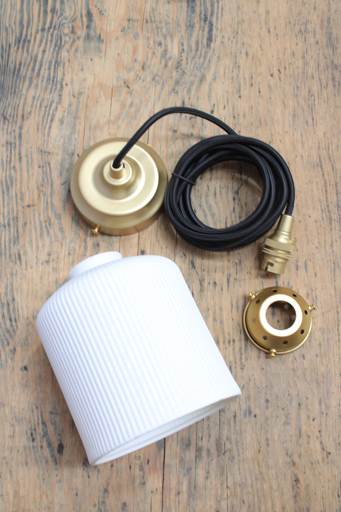 Ribbed ceramic pendant light with black cord and gold/brass hardware, uninstalled on a timber bench. 