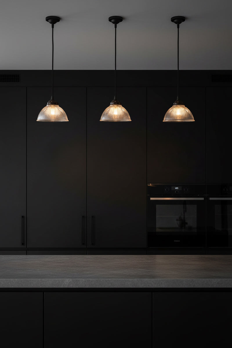 Three como clear pendant lights hanging above a kitchen counter with a dark cupobards. 