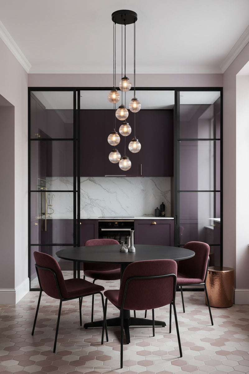 A modern dining room setup featuring a round black table surrounded by four burgundy chairs under a multi-light pendant chandelier showcasing 9 spheres in clear. The room has a contemporary kitchen in the background 
