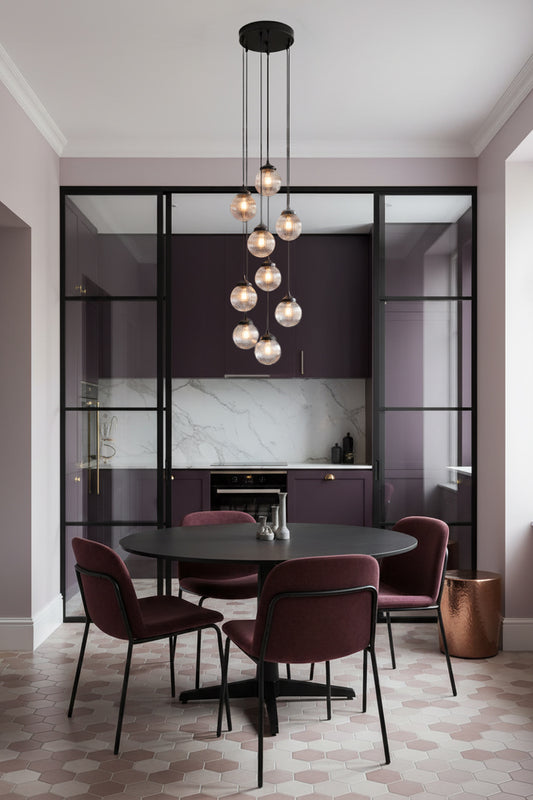 A modern dining room setup featuring a round black table surrounded by four burgundy chairs under a multi-light pendant chandelier showcasing 9 spheres in clear. The room has a contemporary kitchen in the background 