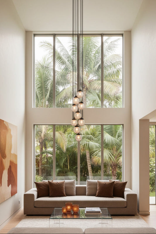A modern multi-light pendant chandelier nine light configurations, with a vintage-inspired design characterised by a picturesque textured pattern hangs in a tall, bright living room with high ceilings, large windows showing palm trees outside. 