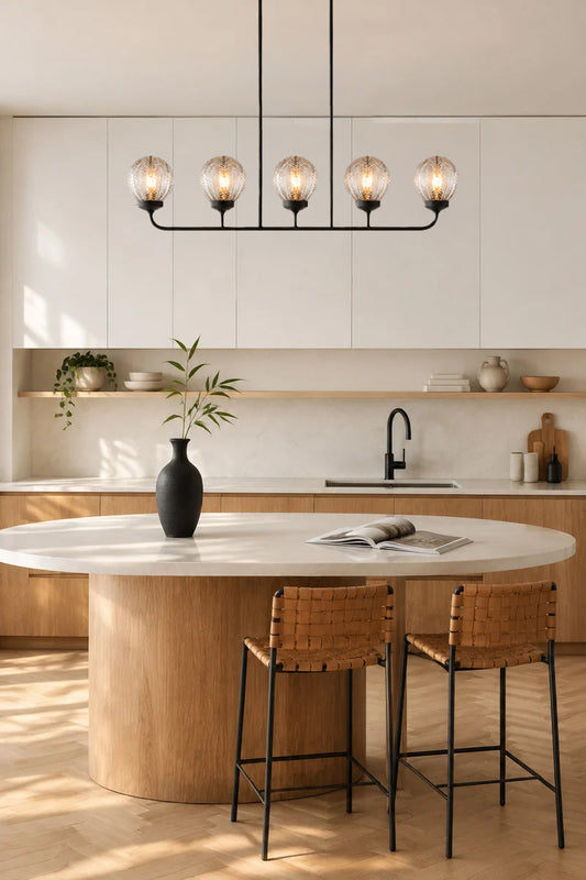 A modern kitchen interior with a round light wood island, white marble top, two woven-back bar stools, a black multi-light pendant with decorative glass shades above the kitchen counter.