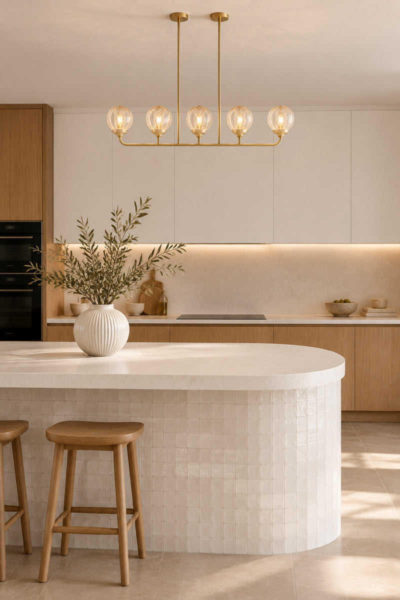 A modern kitchen interior with a curved white island, light wood cabinetry, a gold multi-light pendant chandelier with 5 decorative glass shades over the counter and two wooden bar stools near it.