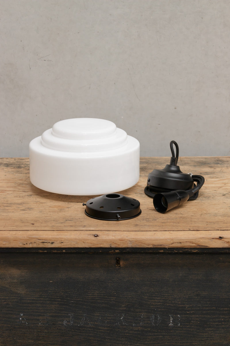 Opal schoolhouse shade with stepped detailing, black pendant cord and black gallery uninstalled on a timber bench. 