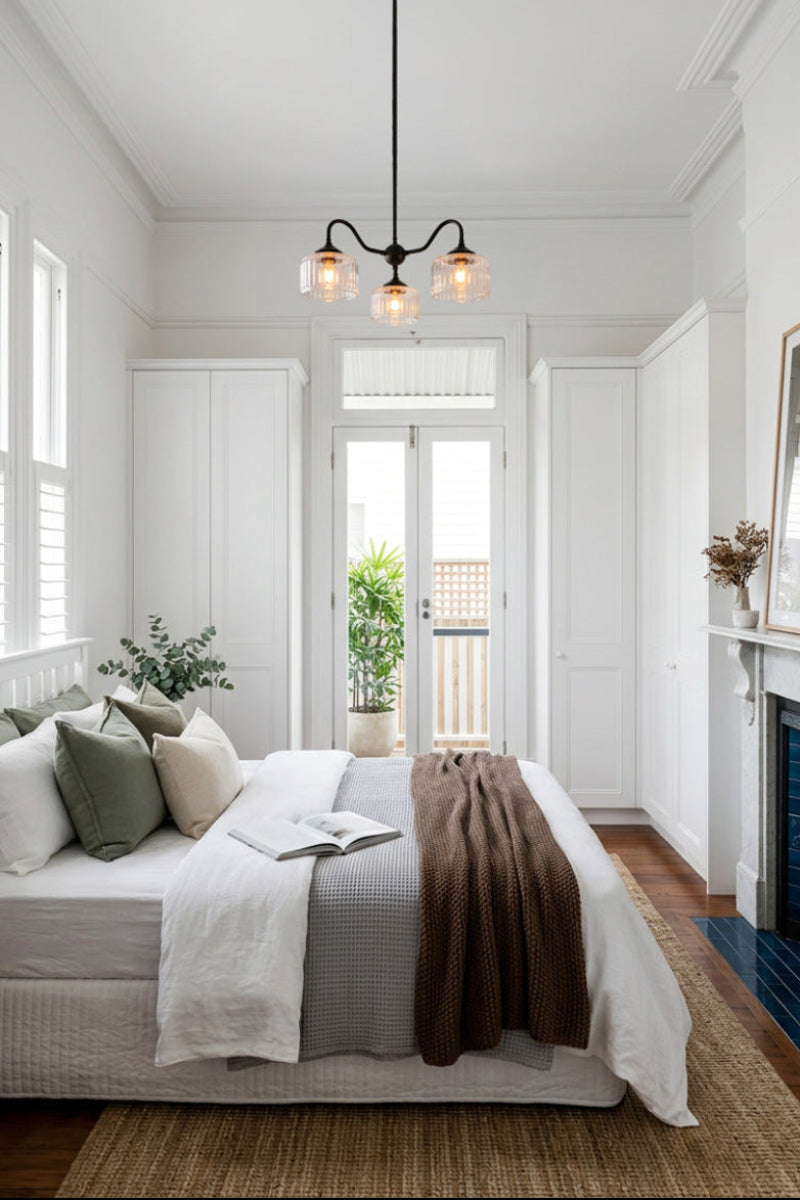 Black three-arm Capri Glass Gooseneck Chandelier with clear fluted glass shades, suspended above a bed in a bright, contemporary bedroom with white panelled walls, timber flooring and neutral furnishings.