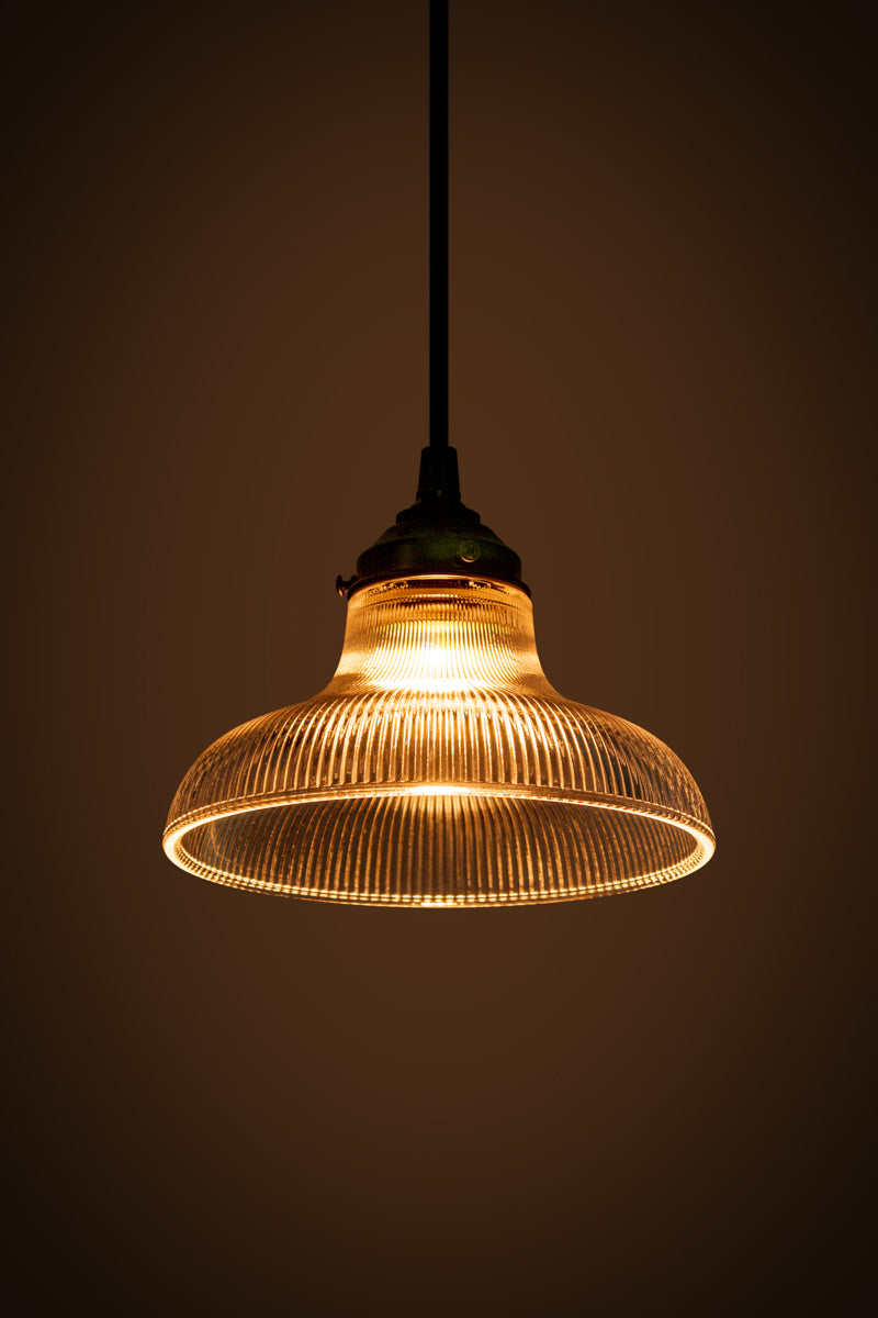 Small clear holohpane glass shade on a black braided cloth cord illuminated in a dark room. 