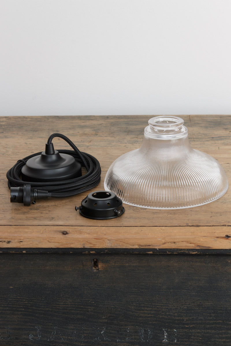 Small clear holohpance glass shade, black braided cloth cord and gallery on a timber bench. 