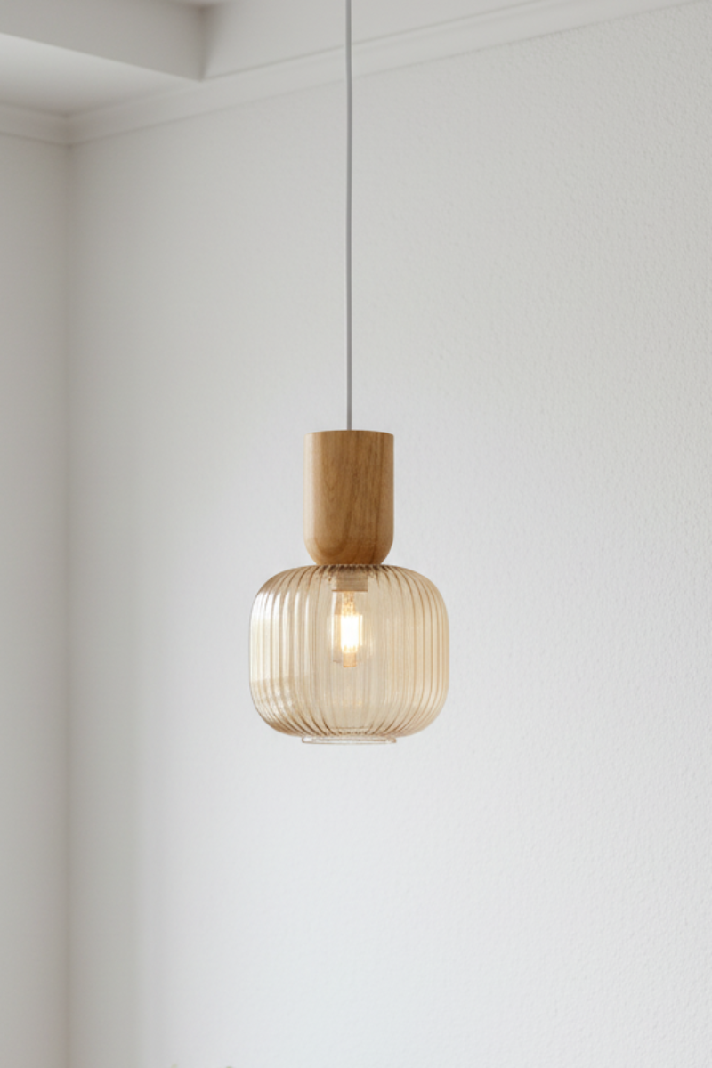 A modern pendant light with a ribbed amber glass shade and a natural wood fitting, hanging in a minimalist white room above a light wood console table with a small potted plant.