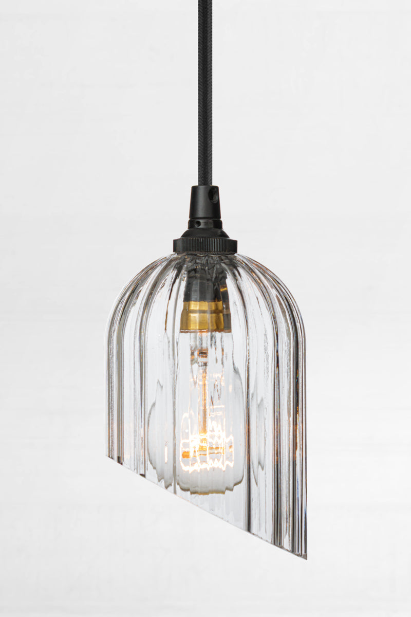 Clear glass elongated shade with fluted detailing and featuring an angle cut, paired with  black pendant cord and clear bulb. 