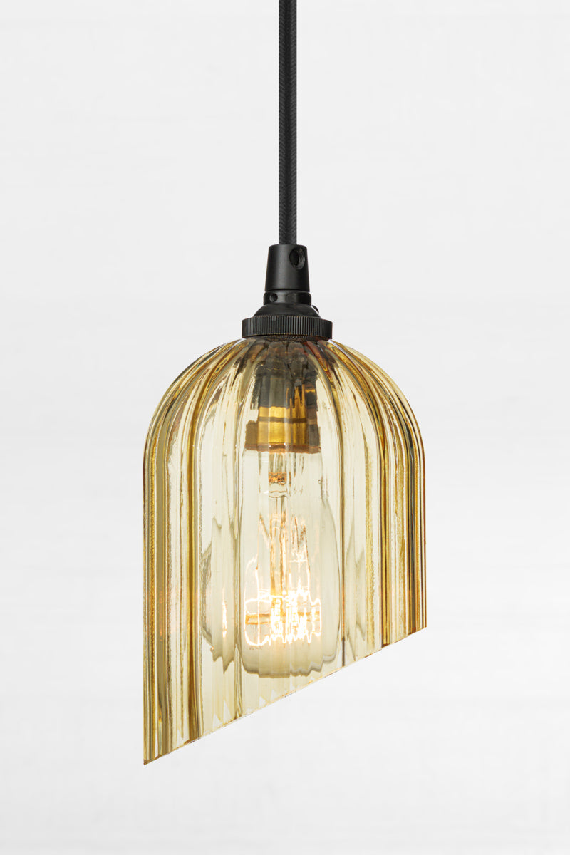 Amber glass elongated shade with fluted detailing and featuring an angle cut, paired with  black pendant cord and clear bulb. 