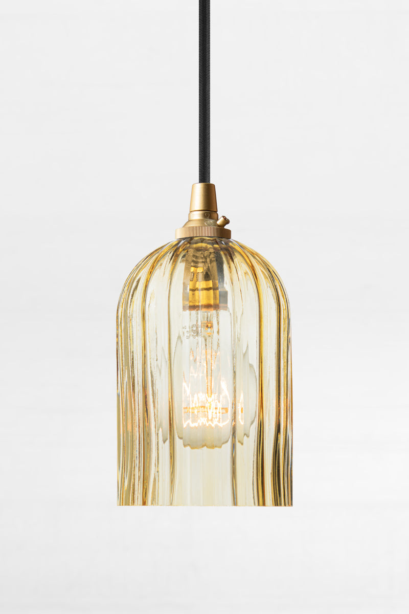 Amber glass elongated shade with fluted detailing, paired with a black pendant cord with gold/brass lampholder and clear bulb. 