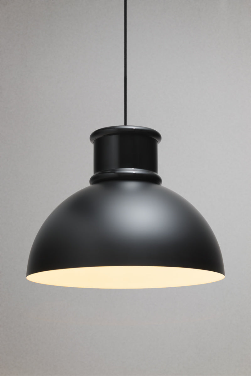 Matt black industrial dome pendant light illuminated with a warm white glow against a neutral background.