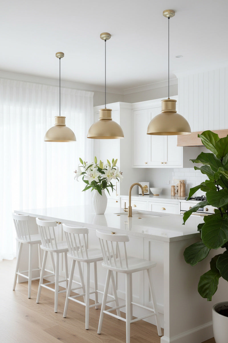 Three gold/brass industrial dome pendant lights hanging above a kitchen island in a bright white kitchen. 