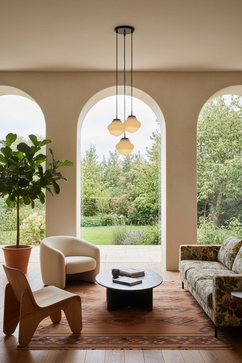 A modern living room interior with arched windows overlooking a green garden. The space includes the Odeon Glass Pendant Light in a 3 drop over the coffee table