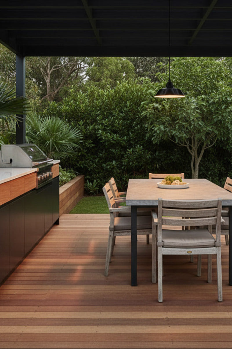 Modern outdoor BBQ and dining area under a pergola, featuring a built-in grill on a counter with wood-look trim, a wooden dining table with a outdoor pendant light over the table