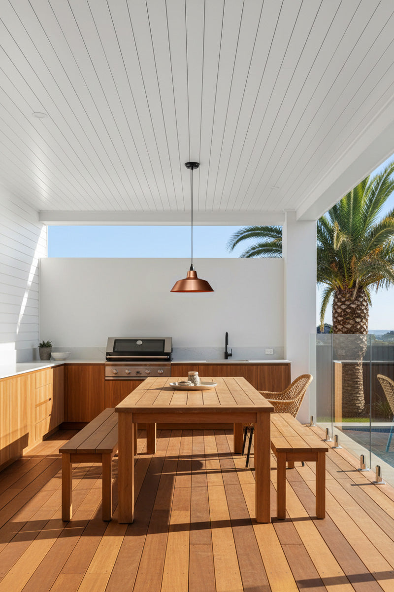 A modern outdoor dining area with a rectangular wooden table and matching benches on a wood deck, under a white slatted ceiling with a copper pendant outdoor light. 
