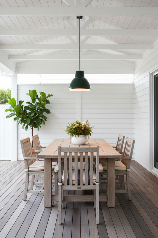 A modern dining set on a covered porch, featuring a Loft Outdoor Green Pendant Light and a wood table and six matching slat-back chairs. 