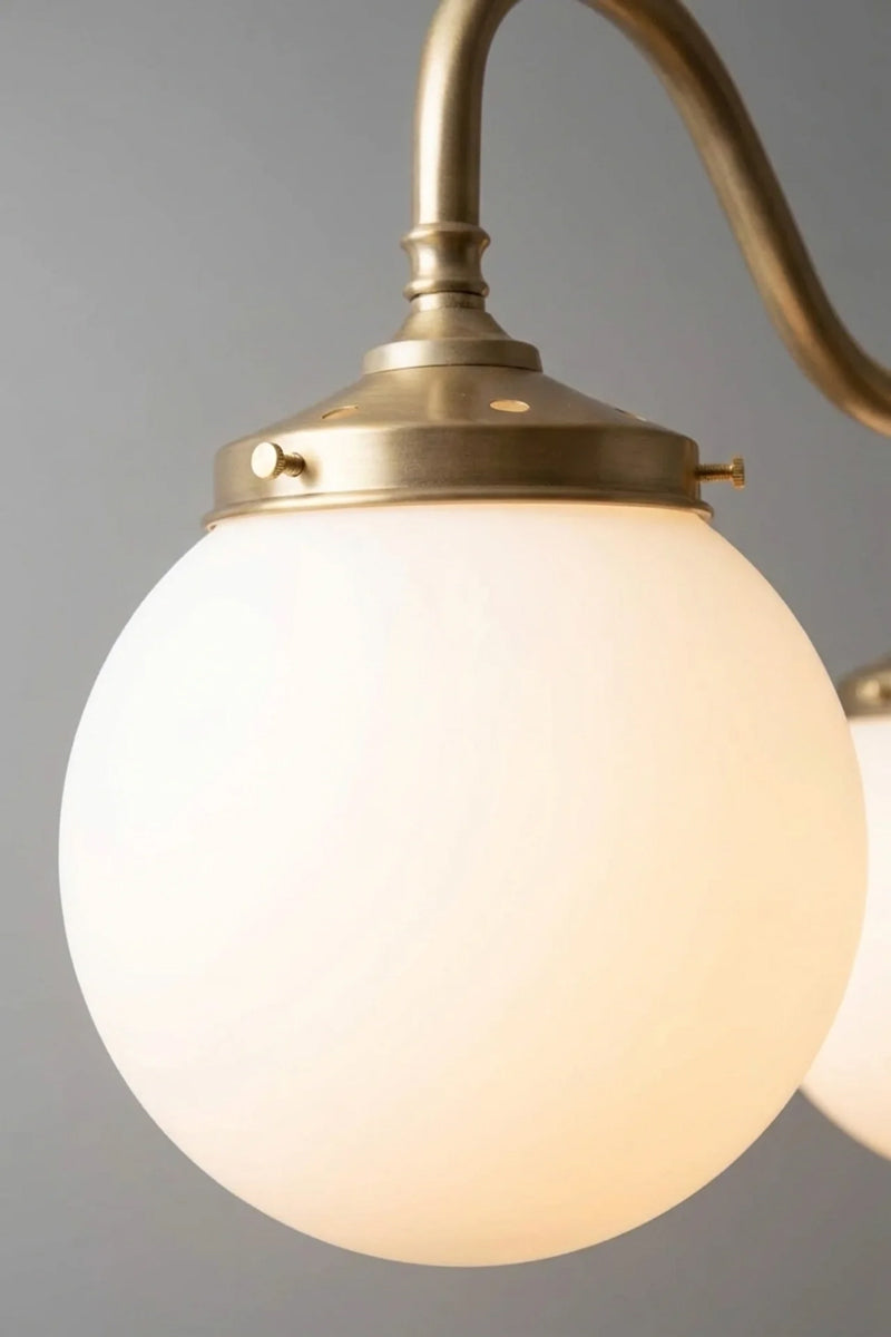 Closeup of small gold three light pendant with opal glass globes on curved arms, lit against a plain grey backdrop