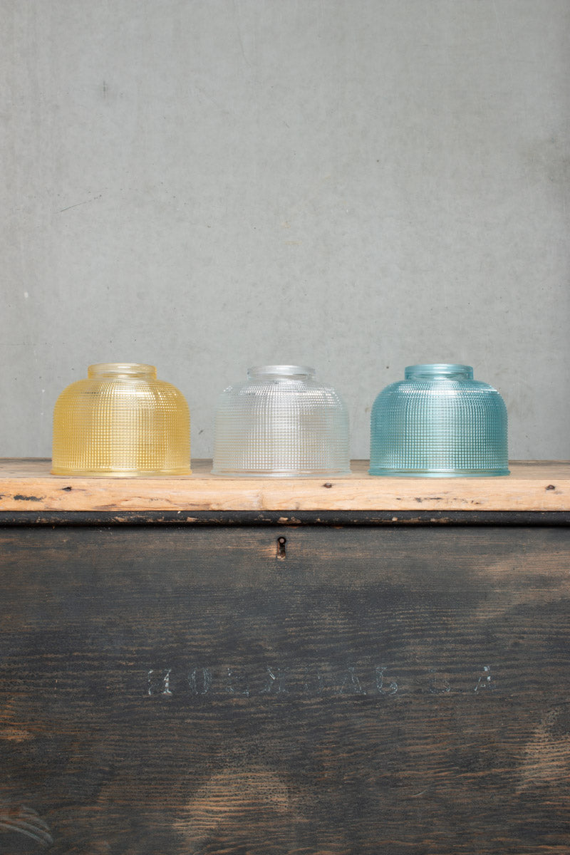 Three small holophane style glass shades in blue, clear and amber, sitting on a timber bench. 