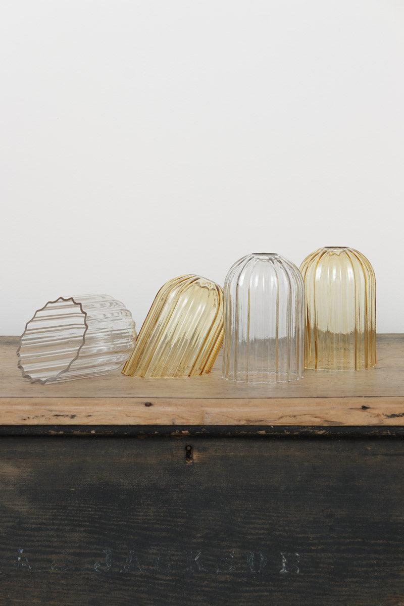 Four glass shades with fluted detailing in amber or clear with a straight or angle cut edge, placed on a timber bench. 