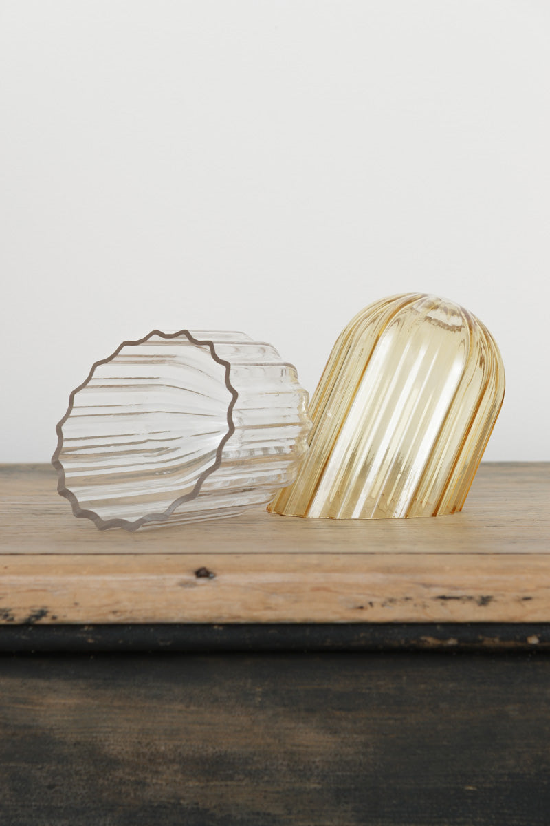 Two glass shades with fluted detailing in amber or clear with an angled cut edge, placed on a timber bench. 