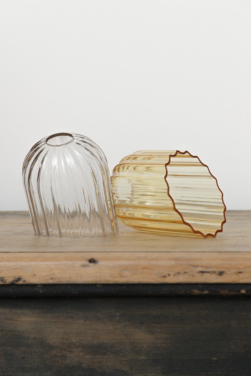 A clear and an amber glass shade with fluted detailing and an angled cut edge, placed on a timber bench. 