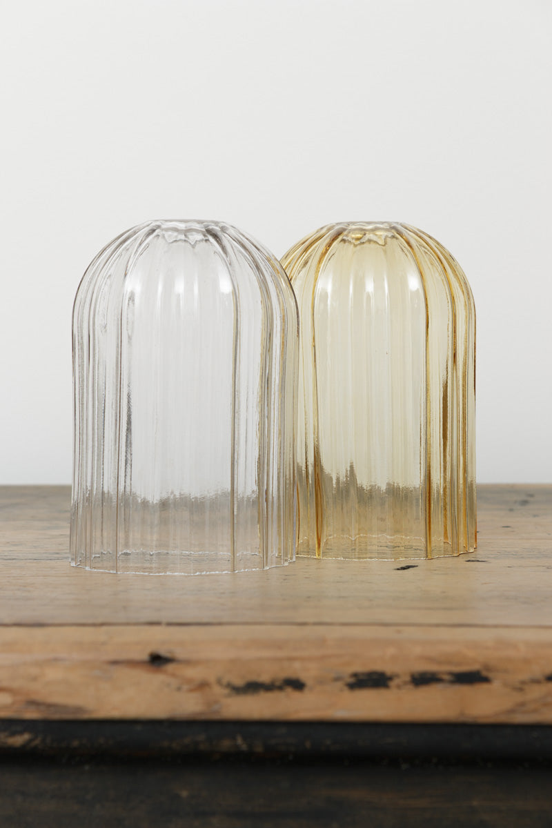 Two glass shades with fluted detailing in amber or clear with an angled cut edge, placed on a timber bench.  