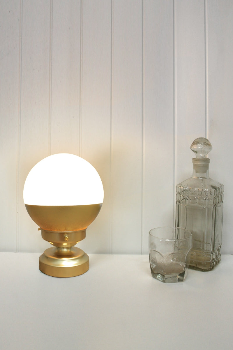 Gold/brass table lam with opal glass shade half painted in coordinating gold/brass in a white spae styled with glassware.