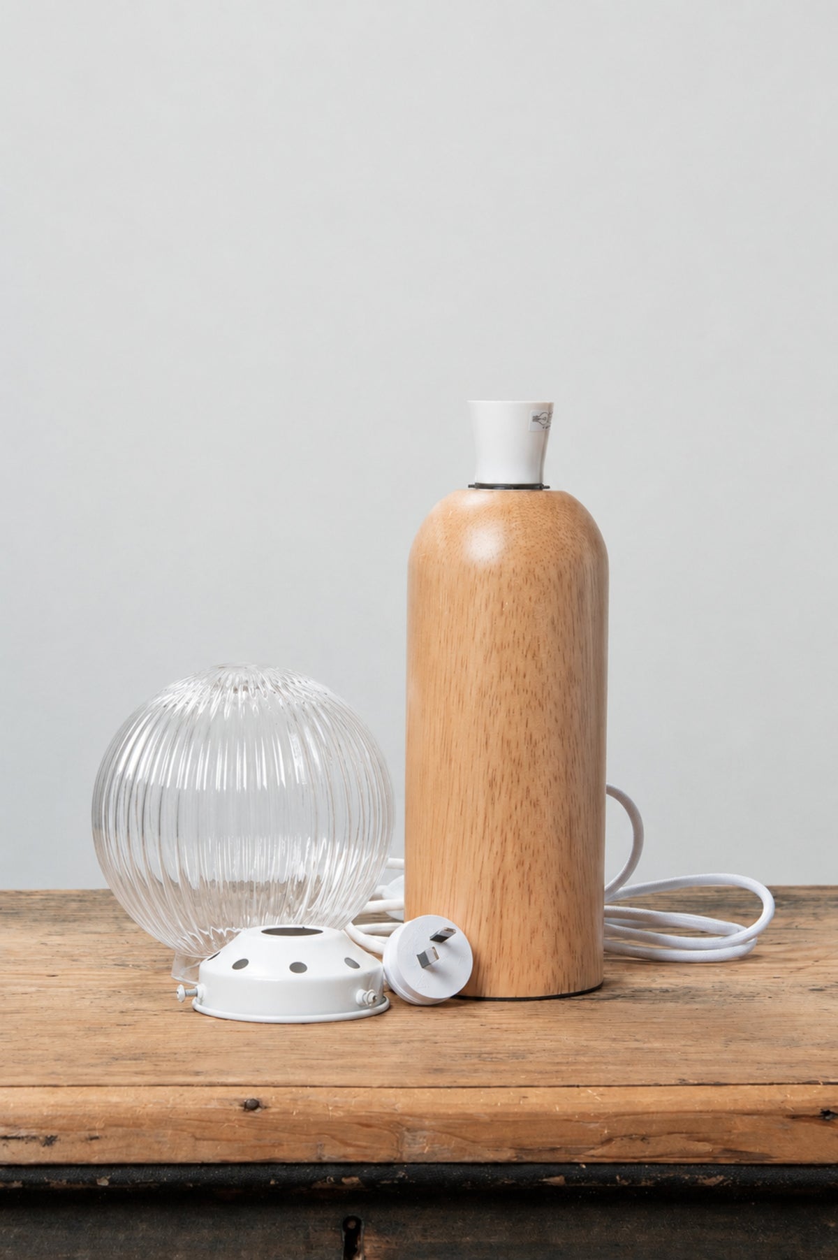 Disassembled table lamp featuring a clear reeded glass ball shade, white gallery and a wood able lamp base on a timber bench. 