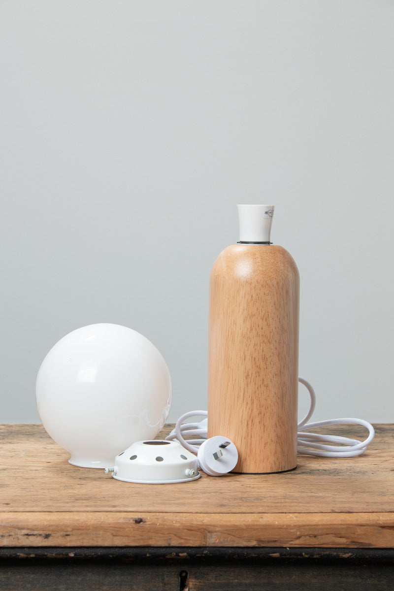 Wood table lamp base with white cord, white metal gallery and opal glass ball shade uninstalled sitting on a timber bench. 