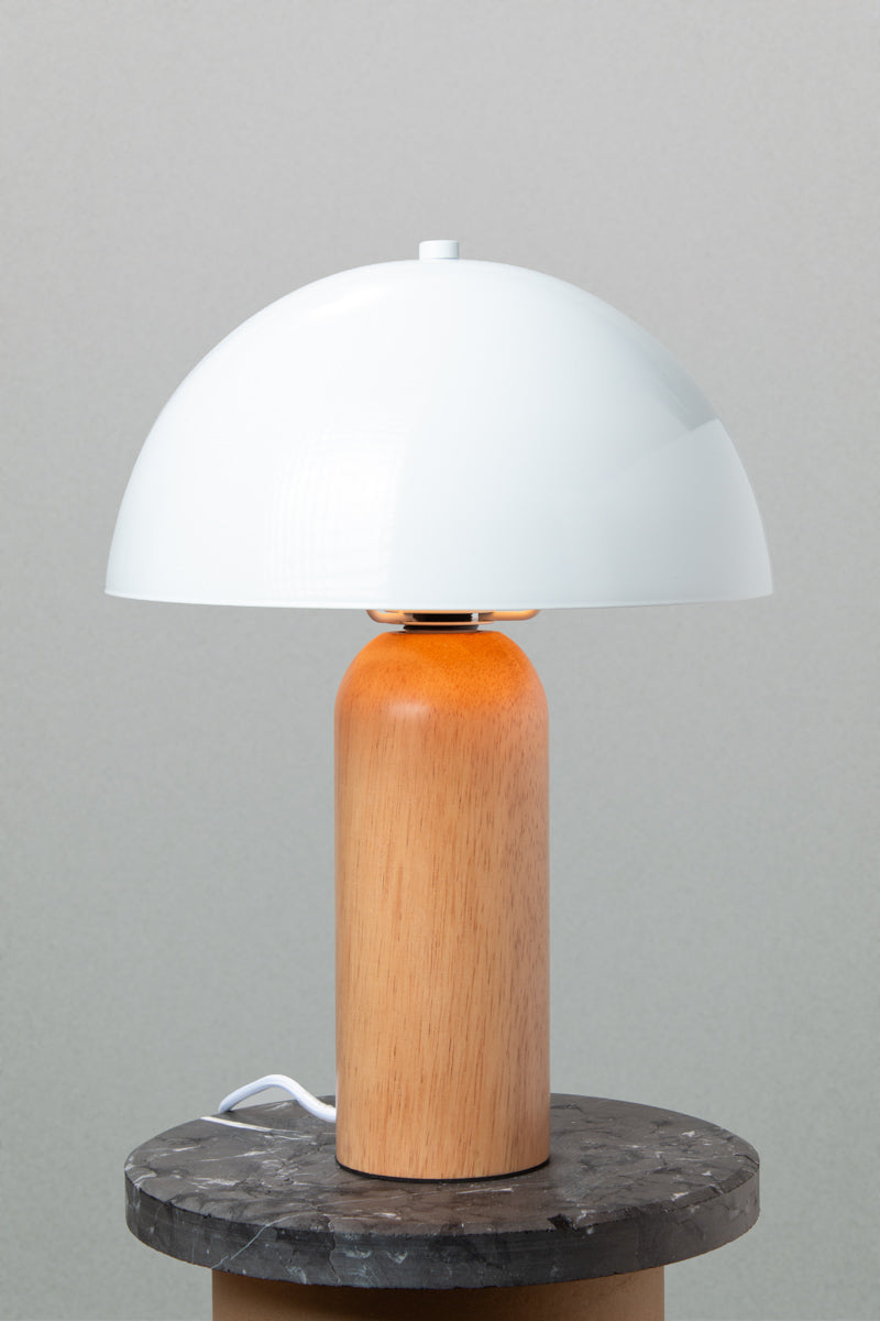 Table lamp with a white metal dome shade and rounded wood base with a natural finish. Lamp placed on a marble look side table. 
