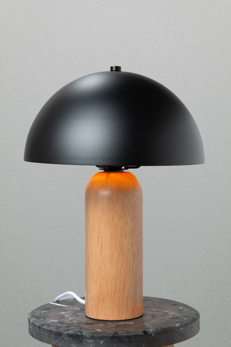 Table lamp with a black metal dome shade and rounded wood base with a natural finish. Lamp placed on a marble look side table. 