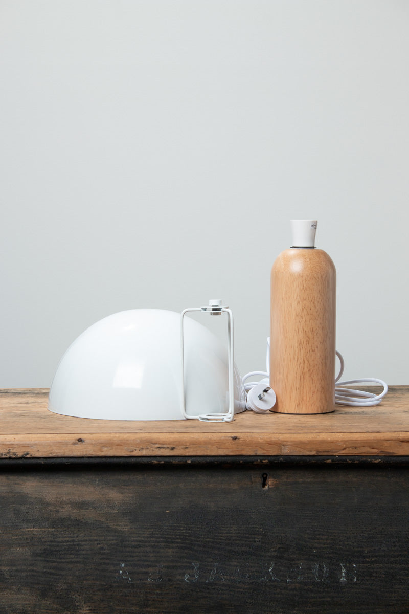 Table lamp with a white metal dome shade and rounded wood base with a natural finish, uninstalled on a timber bench. 