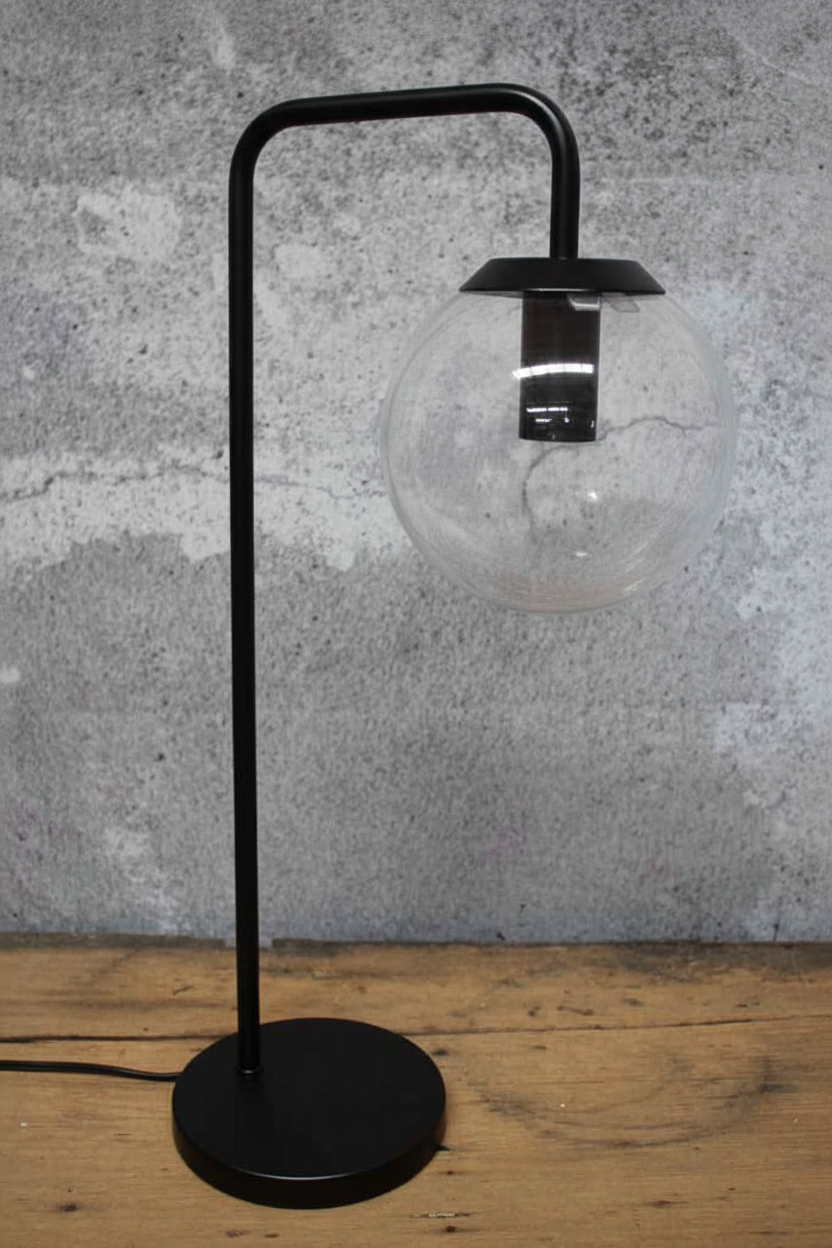 A modern black metal table lamp with a spherical clear glass shade, standing on a round black base against a textured gray wall and wooden tabletop.