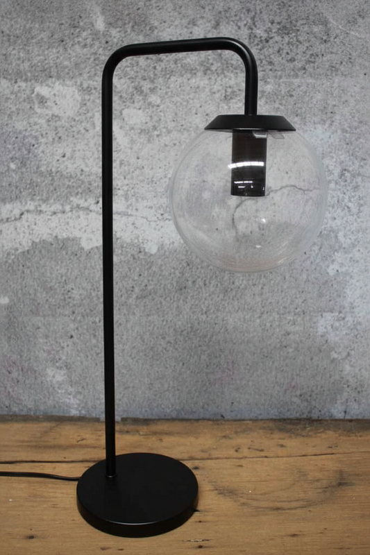 A modern black metal table lamp with a spherical clear glass shade, standing on a round black base against a textured gray wall and wooden tabletop.