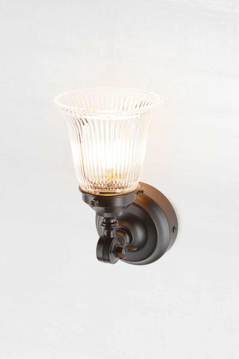 Black wall sconce with adjustable knuckle paired with a small clear glass shade with ribbed detailing. 