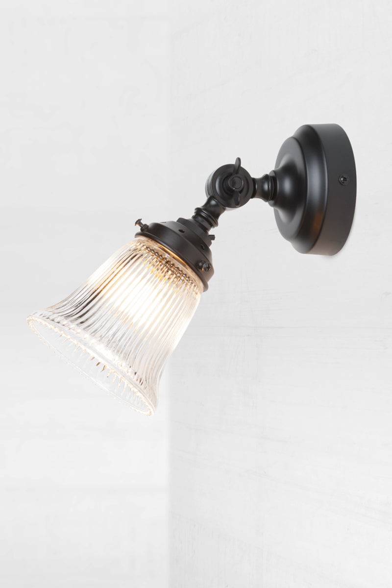 Small clear glass shade with ribbed detailing on a black adjustable batten wall sconce. 