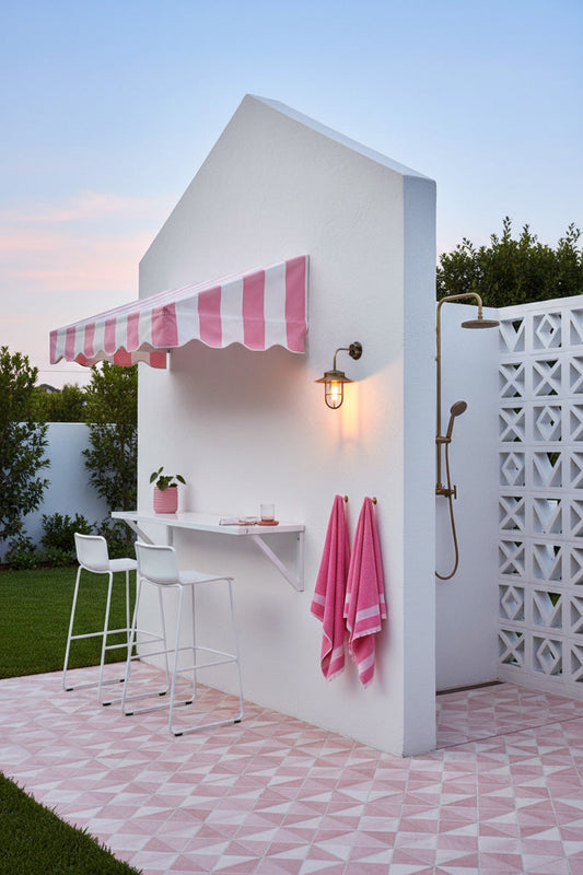 A pink and white patio bar nook with a striped awning and a matching pink-and-white geometric tile floor. A white lattice screen and outdoor shower are visible on the left,  a wall-mounted Solid brass exteriosr light with glass guard, lantern styled light 