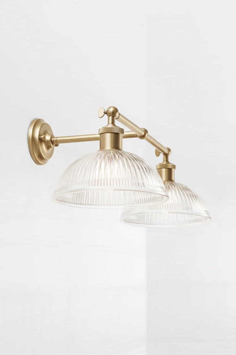 Clear reeded dish shades are mounted to a steel double arm sconce in gold/brass