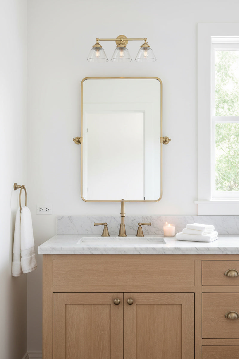 Triple lamp wall sconce in gold/brass with clear shades over a mirror in a classic bathroom. 